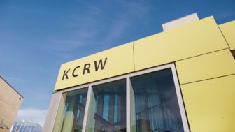 Exterior view of KCRW’s modern studio building in Santa Monica, California, showing its yellow-paneled facade and large glass windows under a clear blue sky.