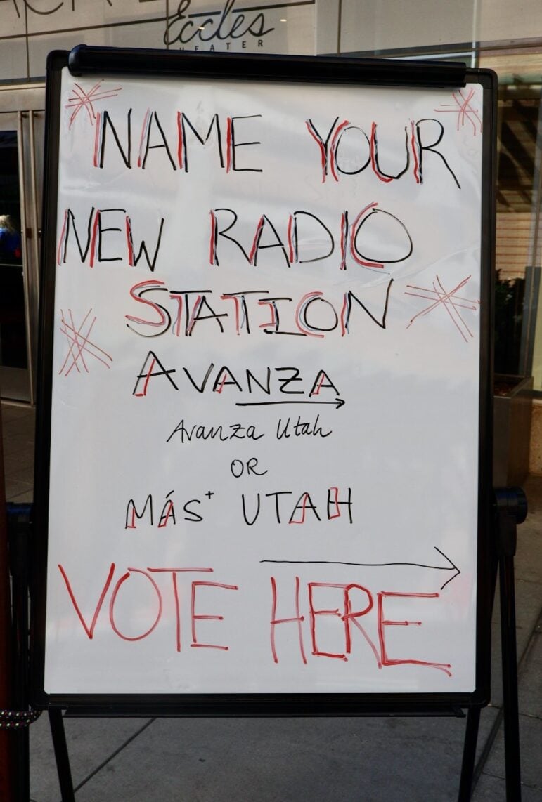 The image shows a whiteboard sign with handwritten text that reads: 

"NAME YOUR NEW RADIO STATION
Avanza
Avanza Utah
or 
Más* Utah

VOTE HERE →"

The text is written in a mix of black and red marker, with decorative stars drawn around the title. The voting choices are "Avanza Utah" and "Más Utah," and there is an arrow pointing to the right next to "VOTE HERE."