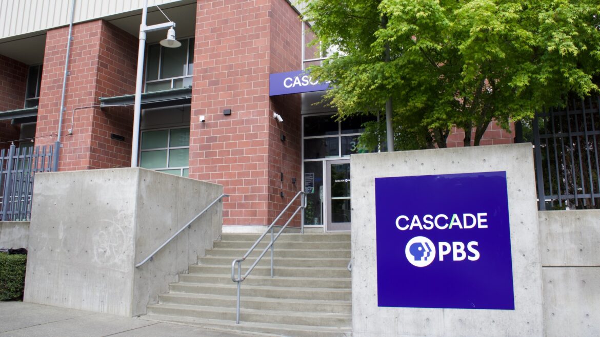 Exterior view of the Cascade PBS building in Seattle, featuring a red brick facade with a concrete staircase leading up to the main entrance. A blue sign displaying the Cascade PBS logo is prominently positioned on the concrete wall to the right of the stairs, with trees and greenery partially visible in the background.