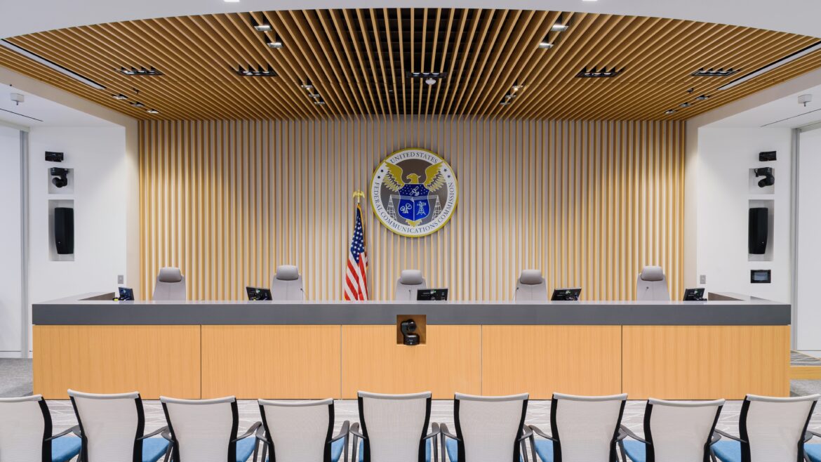 An interior shot of the Federal Communications Commission (FCC) meeting room with a dais, the FCC seal prominently displayed on the wall behind it, an American flag, and chairs arranged for attendees. The room features a modern design with wood paneling and integrated technology.