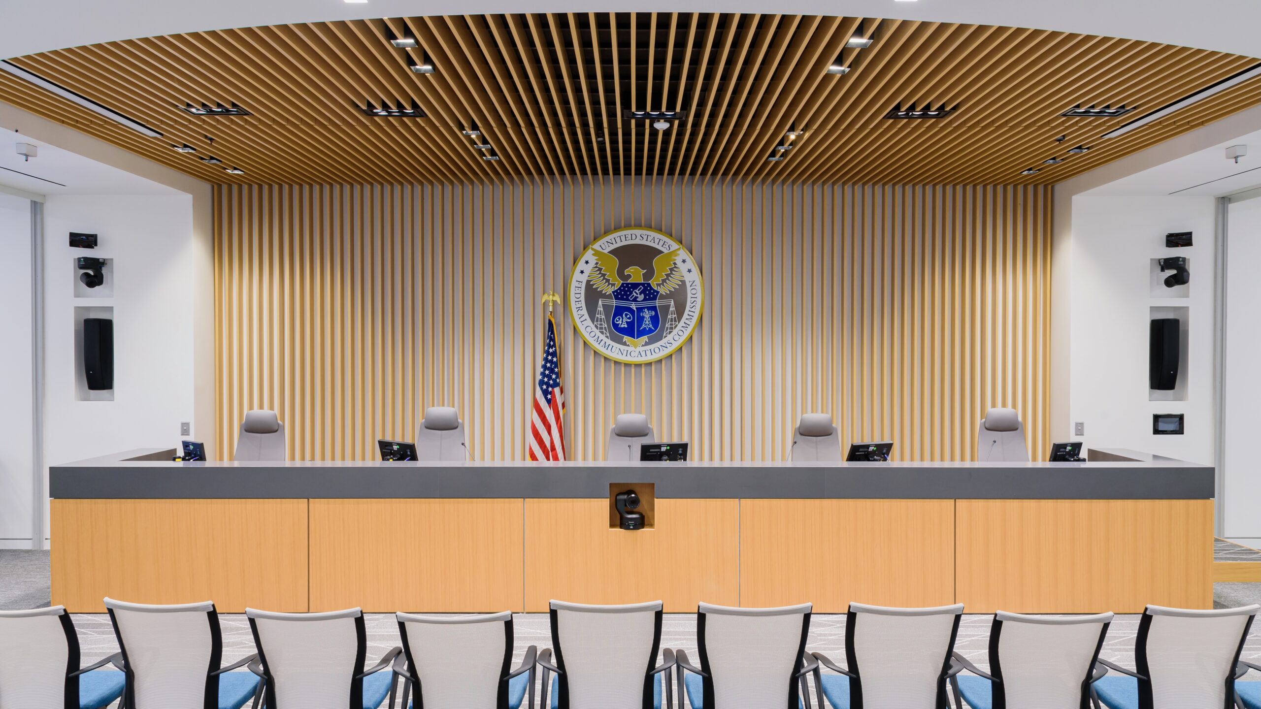 An interior shot of the Federal Communications Commission (FCC) meeting room with a dais, the FCC seal prominently displayed on the wall behind it, an American flag, and chairs arranged for attendees. The room features a modern design with wood paneling and integrated technology.