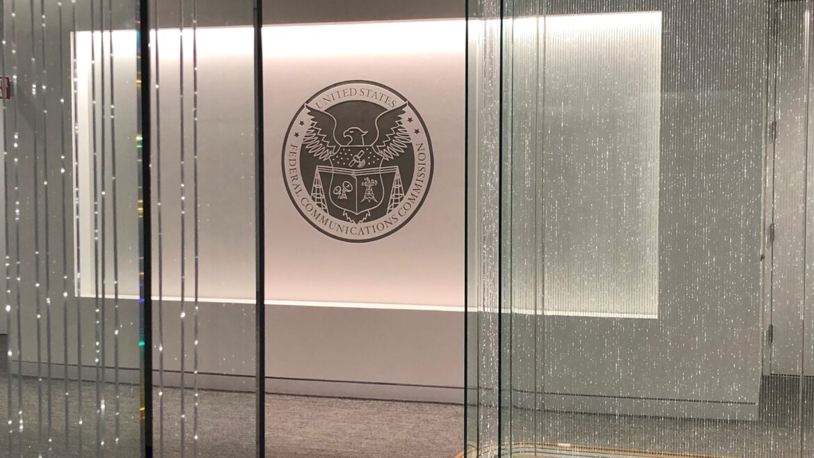 A view of the Federal Communications Commission (FCC) lobby featuring the FCC seal on a white wall, surrounded by glass panels with vertical patterns and soft lighting.