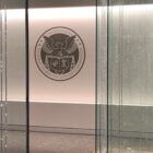 A view of the Federal Communications Commission (FCC) lobby featuring the FCC seal on a white wall, surrounded by glass panels with vertical patterns and soft lighting.