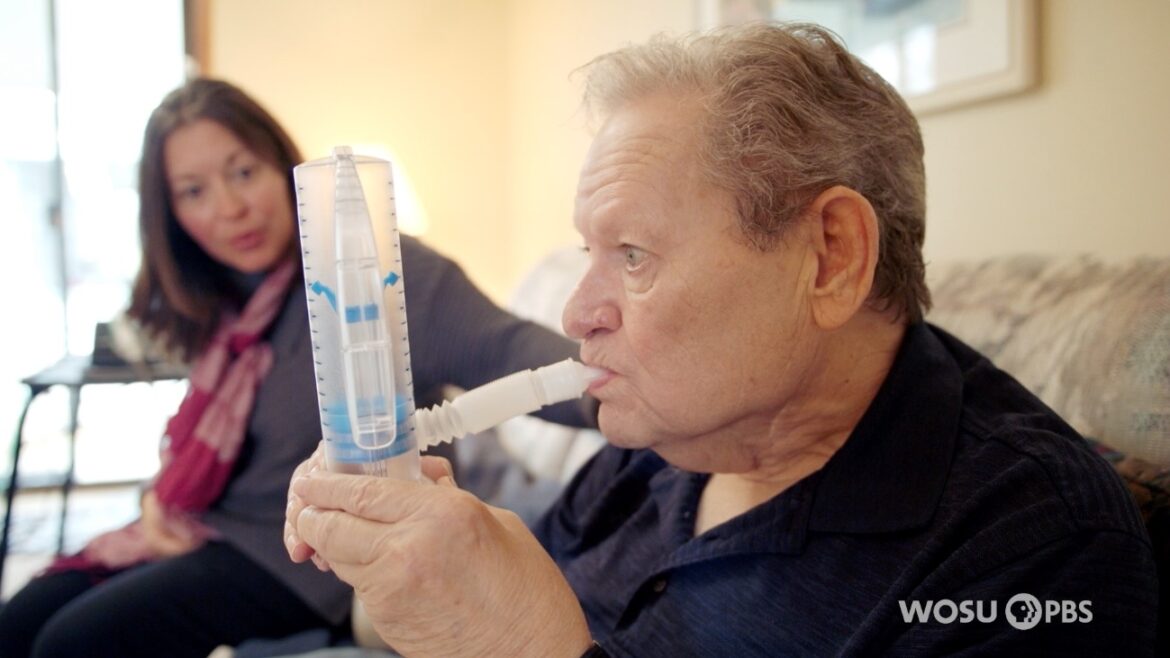 An elderly man measures his breathing with a spirometer while a woman watches.