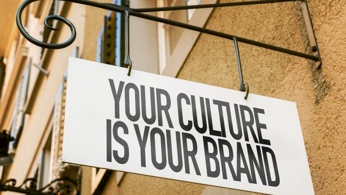 Image of a white sign with black letters that says, "Your Culture is Your Brand," hanging outside of a yellow building