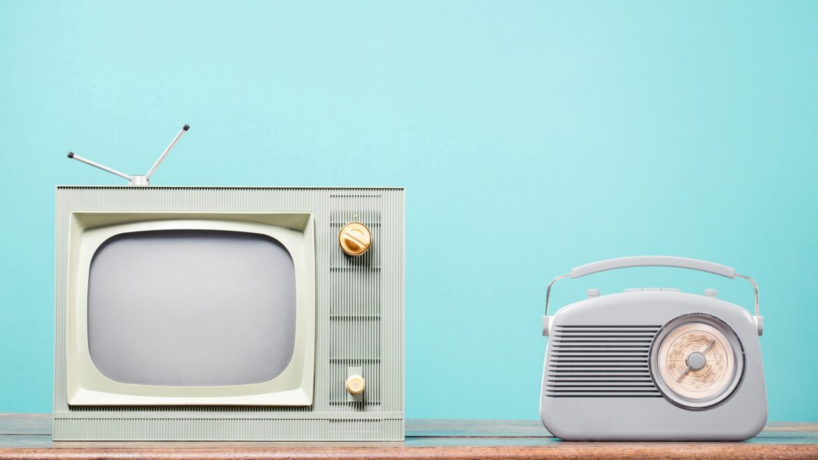 A vintage green television with an antenna and golden knobs sits beside a retro-style gray radio on a wooden surface. The background is a solid light turquoise color, emphasizing the simplicity and nostalgia of the broadcasting equipment.