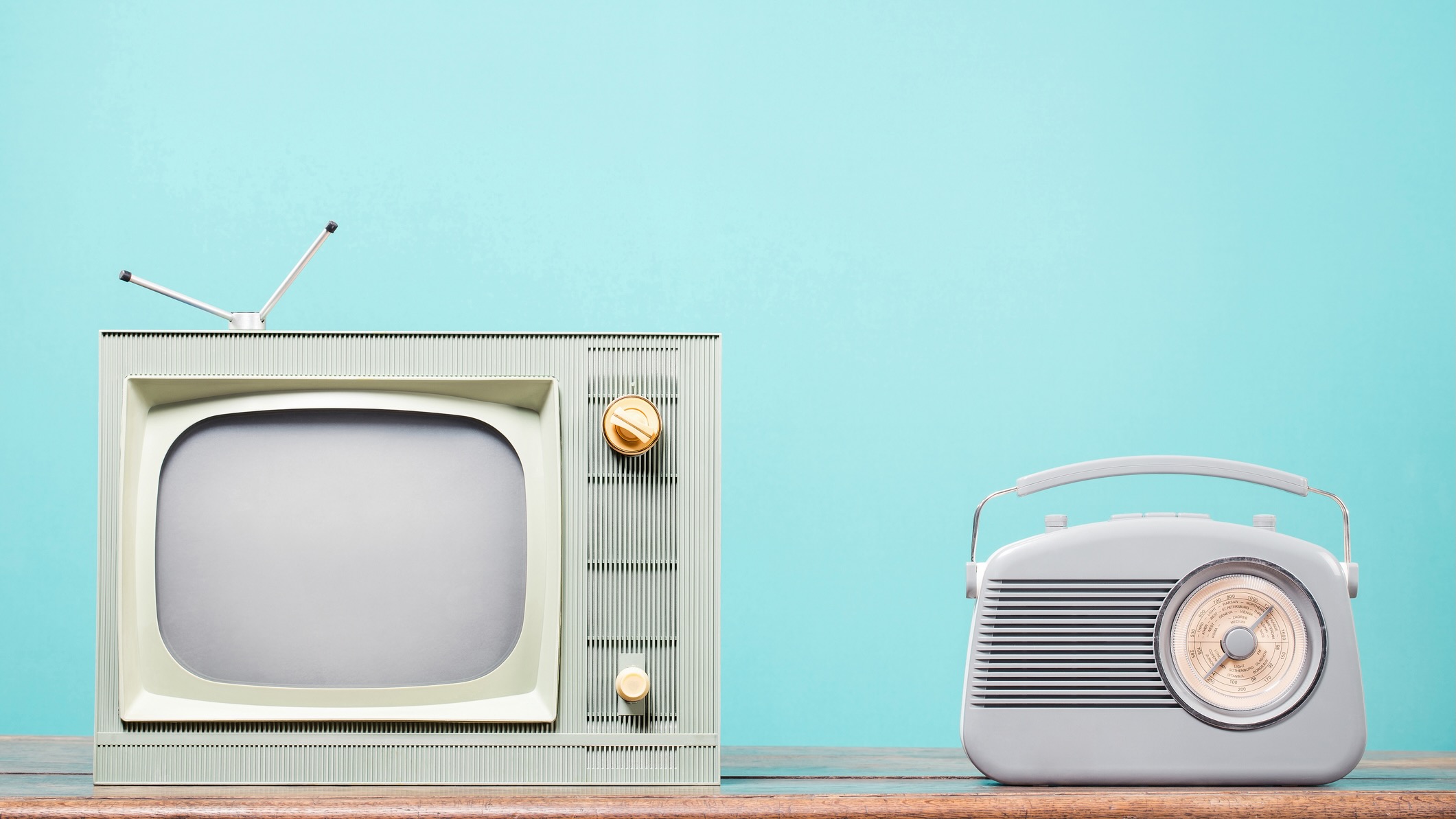 A vintage green television with an antenna and golden knobs sits beside a retro-style gray radio on a wooden surface. The background is a solid light turquoise color, emphasizing the simplicity and nostalgia of the broadcasting equipment.