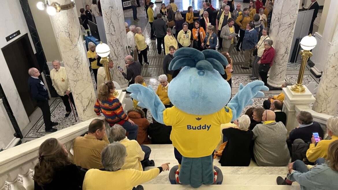 South Dakota Public Broadcasting’s mascot, Buddy, waves to a crowd of supporters gathered on Feb. 7, 2025, at the Capitol Rotunda in Pierre.