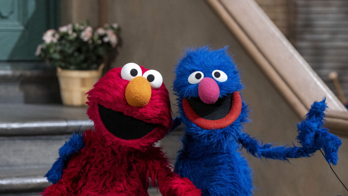 Elmo, a red puppet with large round white eyes, black pupils, and an orange nose, sits on the left, smiling cheerfully. Grover, a blue puppet with similar eyes, a pink nose, and an open-mouthed grin, sits on the right, appearing excited. They are posing together on the steps of a building, leaning toward each other in a friendly manner. A potted plant with pink flowers is visible in the background.