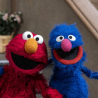 Elmo, a red puppet with large round white eyes, black pupils, and an orange nose, sits on the left, smiling cheerfully. Grover, a blue puppet with similar eyes, a pink nose, and an open-mouthed grin, sits on the right, appearing excited. They are posing together on the steps of a building, leaning toward each other in a friendly manner. A potted plant with pink flowers is visible in the background.