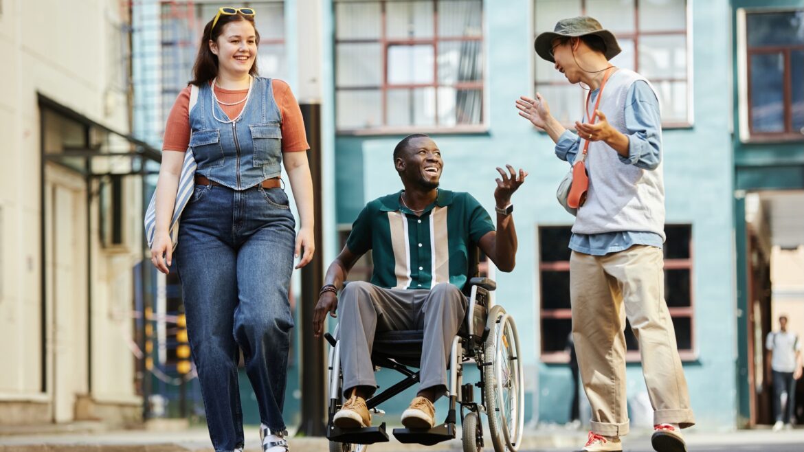 Colorful front view portrait of diverse group of friends with African American man using wheelchair moving towards camera in urban city setting and chatting cheerfully
