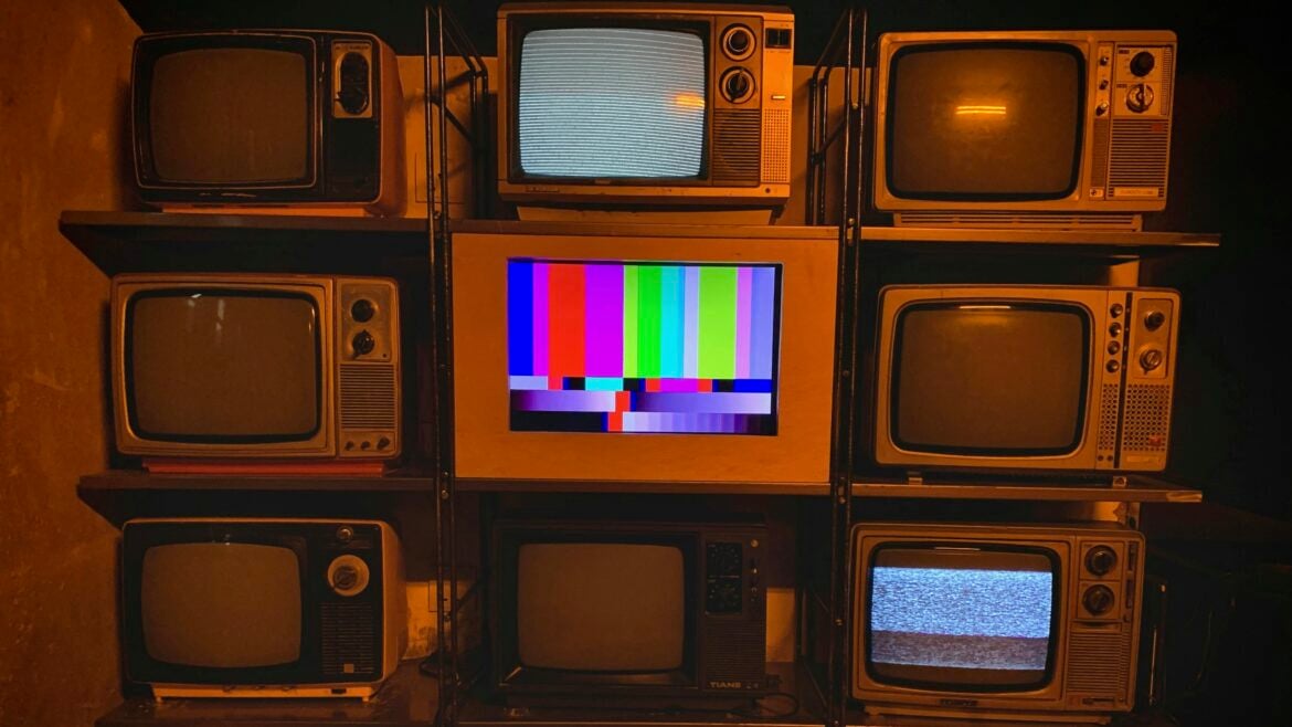 A wall of vintage television sets, most turned off or showing static, with one in the center displaying a colorful test pattern, evoking themes of broadcast disruption or decline.