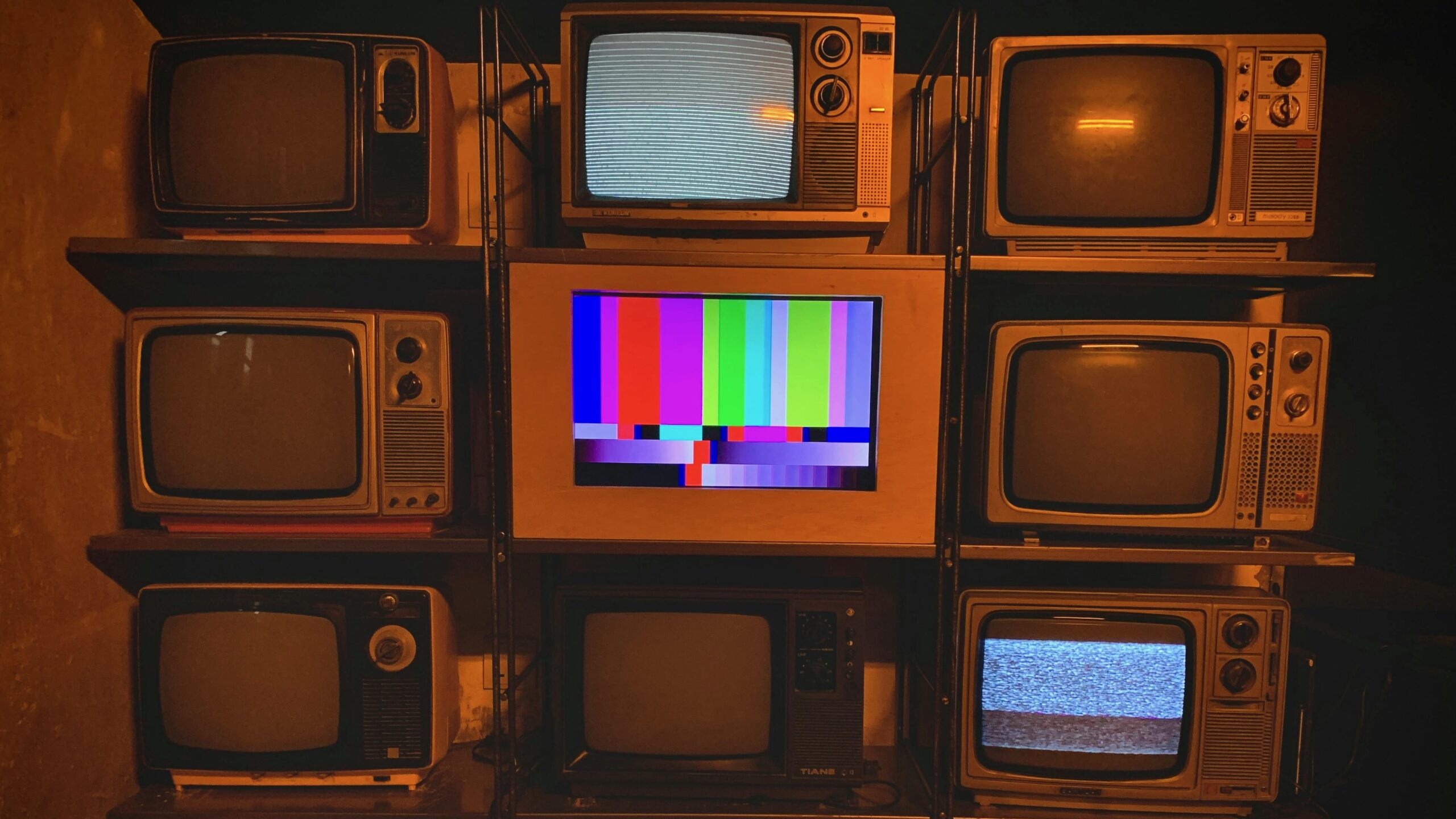 A wall of vintage television sets, most turned off or showing static, with one in the center displaying a colorful test pattern, evoking themes of broadcast disruption or decline.