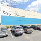 Exterior view of a South Carolina ETV and Public Radio building, showing a large blue and white facade with the SCETV and Public Radio logos displayed. Several cars are parked in the lot in front of the building, and the surrounding area features palm trees and a clear sky.