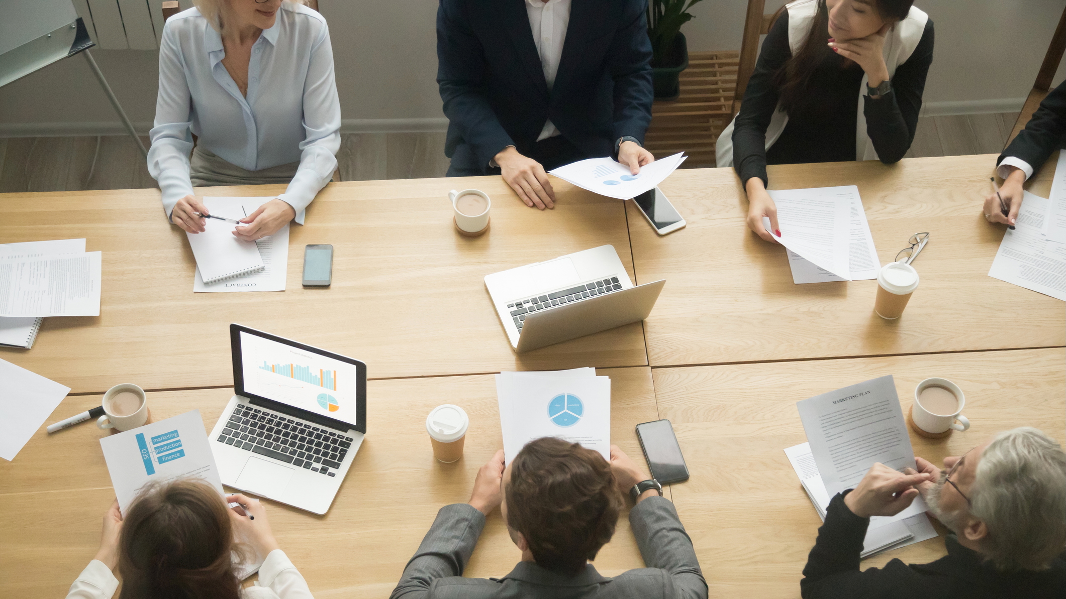 Alt text: A diverse group of professionals sits around a large wooden conference table in a modern office, engaged in a business meeting. Laptops, printed charts, documents, coffee cups, and smartphones are scattered on the table, suggesting a discussion of strategy. The image is viewed from above.