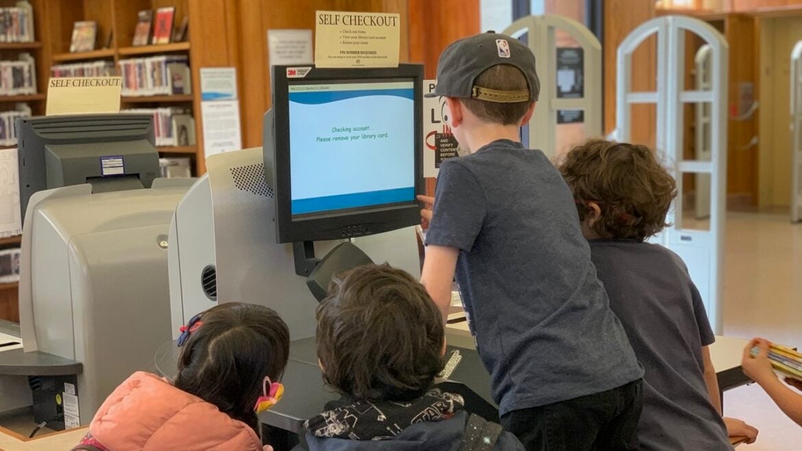 A group of young children crowds around a self-checkout station in a library. One child interacts with the monitor, which displays a message about removing a library card. The other children watch closely. Bookshelves and security gates are visible in the background.