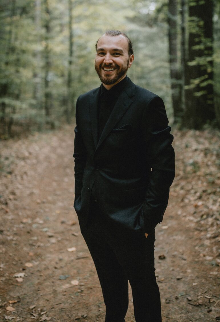 Joshua Rae stands smiling in a forest path, dressed in a dark suit with hands in his pockets. The background is filled with tall trees and soft natural light.