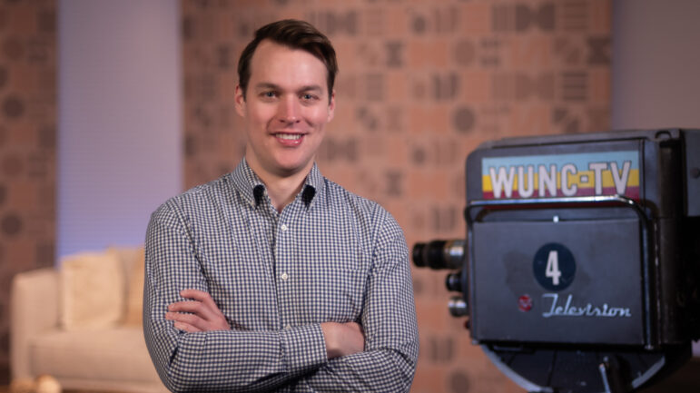 Josh Clinard stands smiling with arms crossed in a studio setting, next to a vintage WUNC-TV camera. A patterned wall and sofa are visible in the background.