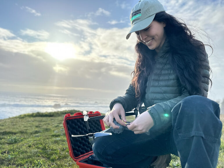 Hannah Lee kneels on a grassy bluff near the ocean, smiling while preparing a drone. She wears a green jacket and a Northern California Public Media cap, with a red equipment case open beside her.