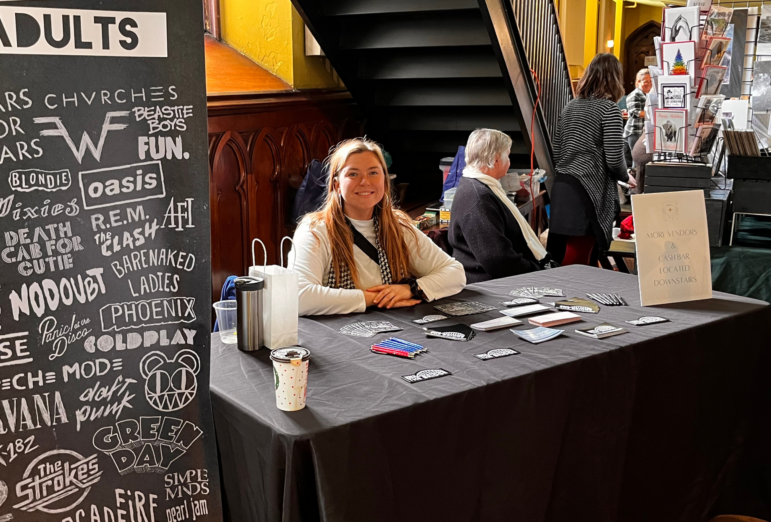 Elyse Breeze sits at a vendor table covered in merchandise, smiling at the camera during an indoor market event. A large black display board next to her lists band names in white text, and a sign nearby points to more vendors and a cash bar downstairs.