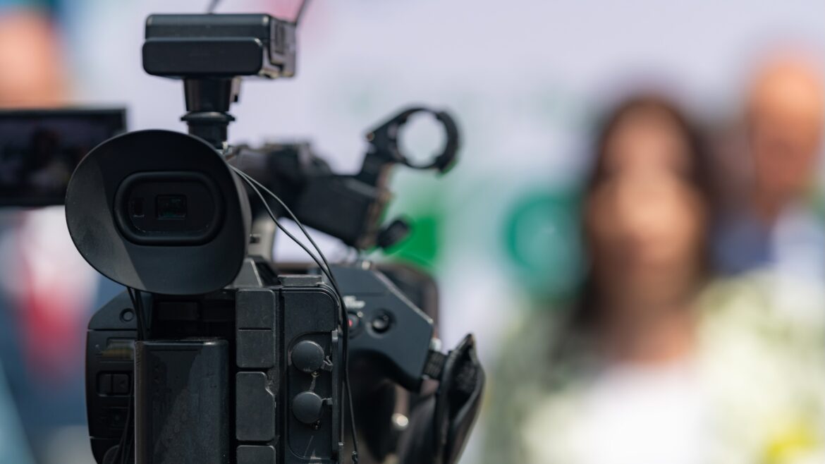 Close-up of a professional video camera recording an interview, with the camera in sharp focus and a blurred figure of a woman in the background.