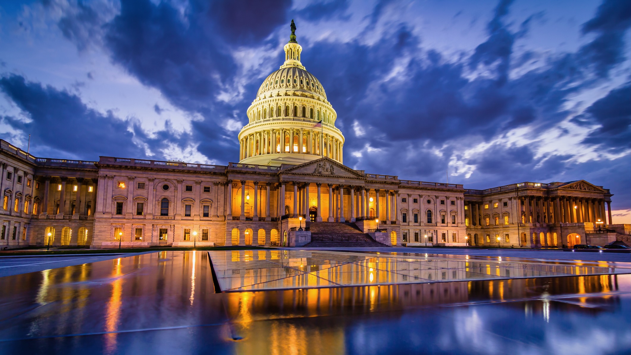 The U.S. Capitol building illuminated at dusk, with dramatic storm clouds overhead and reflections of its lights on a glassy surface in the foreground.