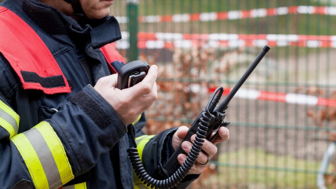 A firefighter in uniform holds and operates a handheld two-way radio, with caution tape and a fence in the background.