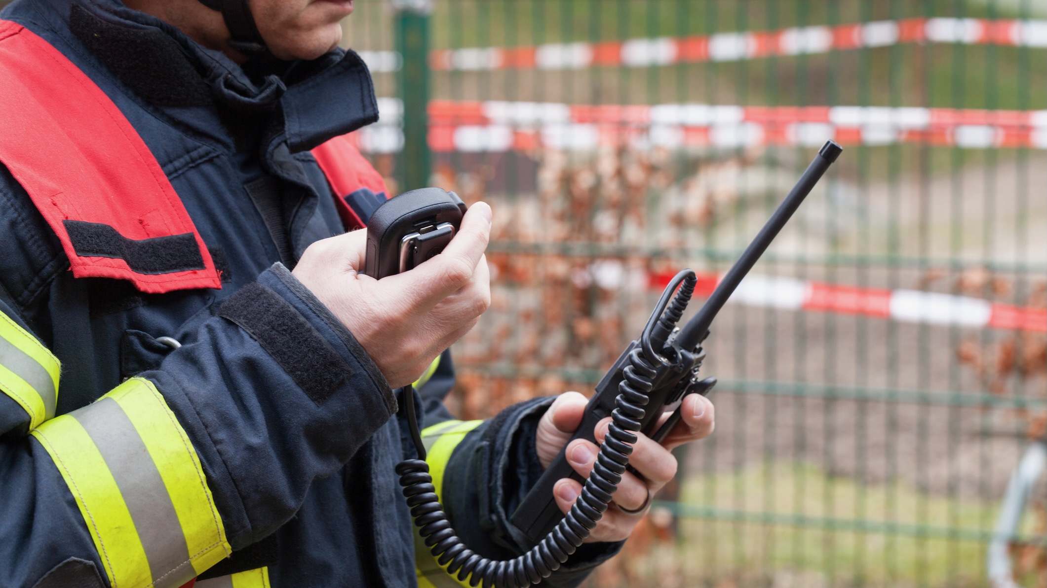 A firefighter in uniform holds and operates a handheld two-way radio, with caution tape and a fence in the background.