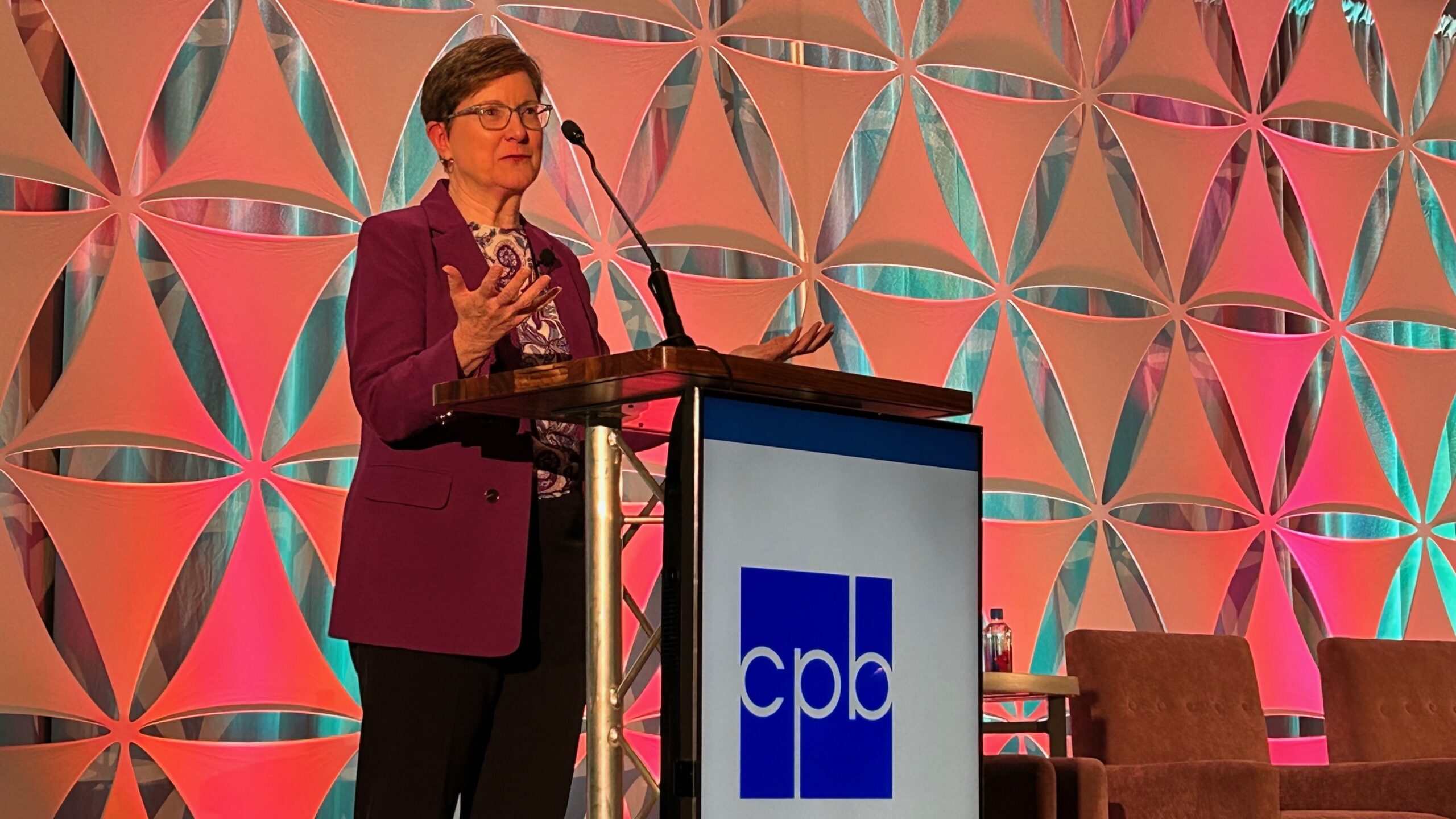 CPB COO Kathy Merritt speaks at a podium with a microphone Thursday at the Public Media Business Association conference. She is wearing a purple blazer and glasses, gesturing with her hands. The podium displays the CPB logo, and the backdrop features a geometric pattern lit with pink and teal lighting.