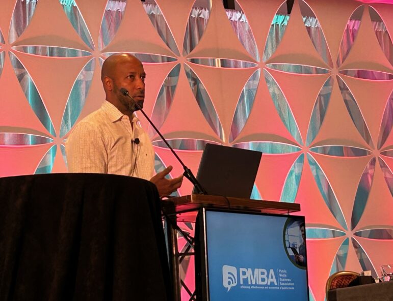Moustapha Abdul, CPB's senior director of station and system analysis, speaks at a podium with a microphone headset during the 2025 PMBA conference. Behind him is a geometric backdrop lit with pink and teal lighting. The podium displays the PMBA (Public Media Business Association) logo.