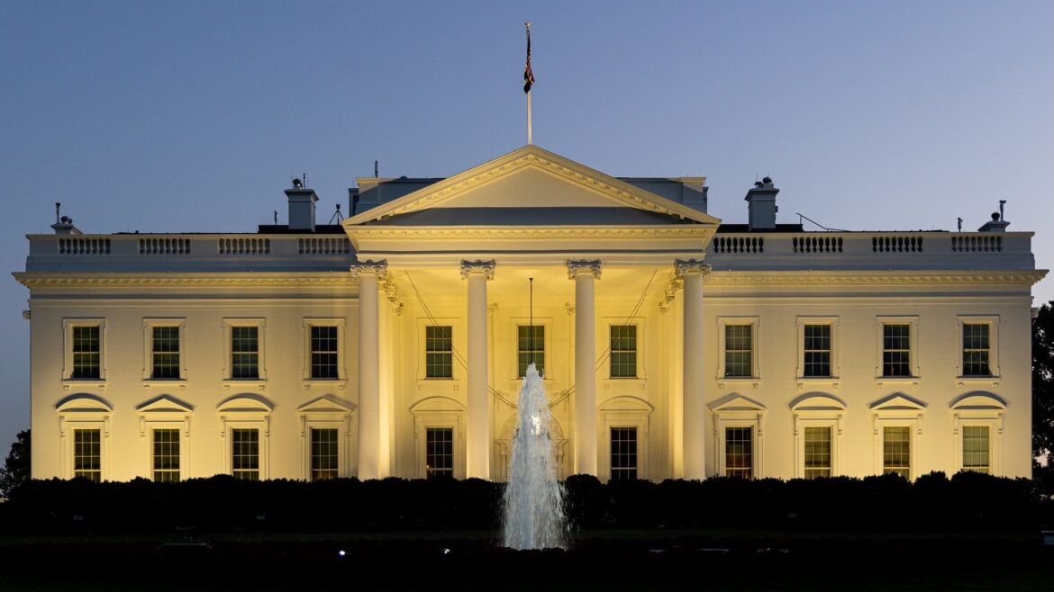 The White House at dusk, illuminated by exterior lights that highlight its white facade and columns. A fountain is centered in the foreground, spraying water upward. The sky is clear and fading into evening blue.