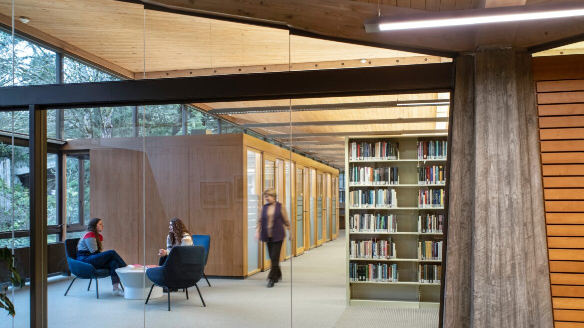 Interior view of the Boley Law Library at Lewis & Clark Law School, showing a modern reading area with two people seated and talking at a small round table, a person walking past wooden study rooms, and bookshelves filled with books. Large windows and a wood-paneled ceiling bring in natural light and views of trees outside.