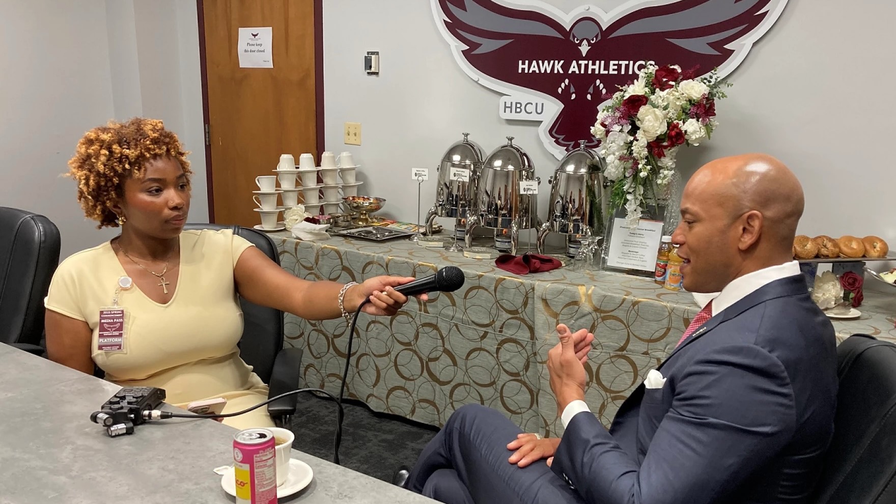 University of Maryland Eastern Shore student journalist Sydney Rutledge-Smith interviews Maryland Governor Wes Moore, holding a microphone toward him as they sit across a table in a room with a buffet setup and a “Hawk Athletics” logo on the wall.