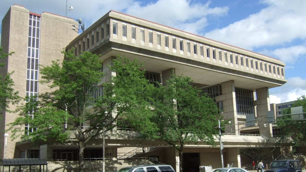 Exterior view of Vilas Communication Hall at the University of Wisconsin–Madison, a large concrete and brick academic building with a cantilevered upper floor and tall vertical windows, partially obscured by leafy trees.