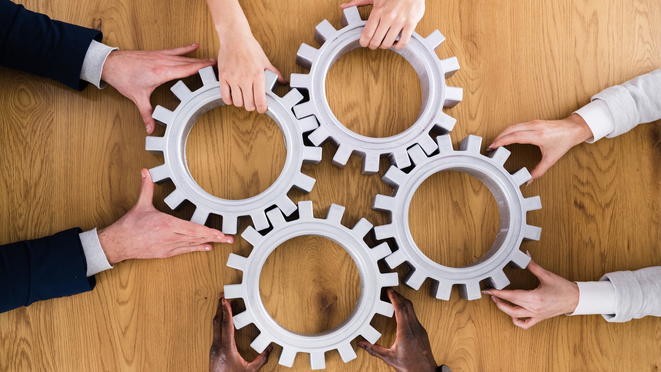 A group of diverse hands working together to connect five interlocking gear-shaped pieces on a wooden table, symbolizing teamwork and collaboration.