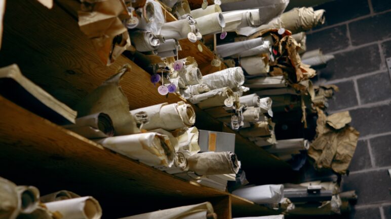 A side view of hundreds of rolled-up maps lining half a dozen wooden shelves. The old canvases are various shades of beige with circular purple or yellow tags hanging at their ends. 