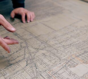 A person's hand gestures across an old, tan-colored, hand-drawn map of mine tunnels under city blocks
