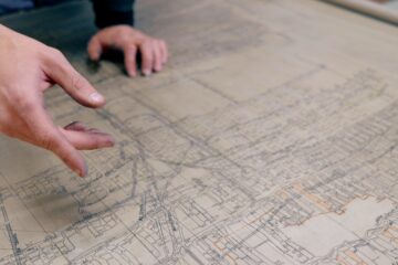 A person's hand gestures across an old, tan-colored, hand-drawn map of mine tunnels under city blocks