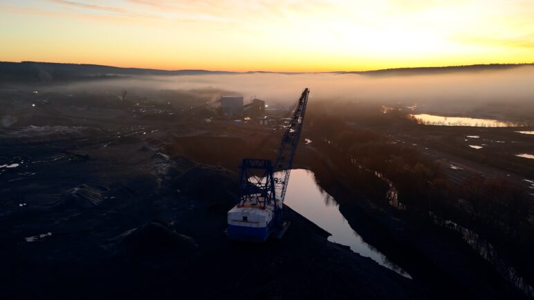 A drone view at dawn, overlooking a surface mining site. A large dark blue crane is in the center, pointed to the right over a small body of water. Haze obscures the mining equipment farther away. 