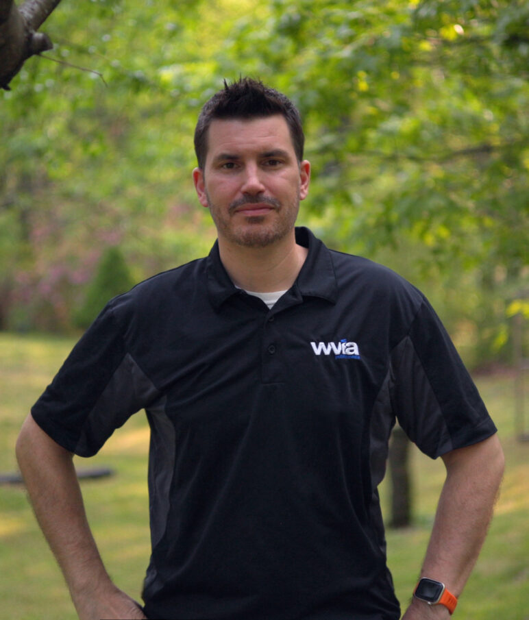 A waist-up portrait of WVIA Multimedia Producer Tim Novotney, a white man with dark brown hair wearing a navy WVIA polo. 