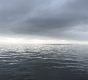 A wide expanse of calm water under a dark, overcast sky. A long, narrow bridge stretches across the horizon in the distance. The scene is moody and serene, with soft ripples on the surface of the water.