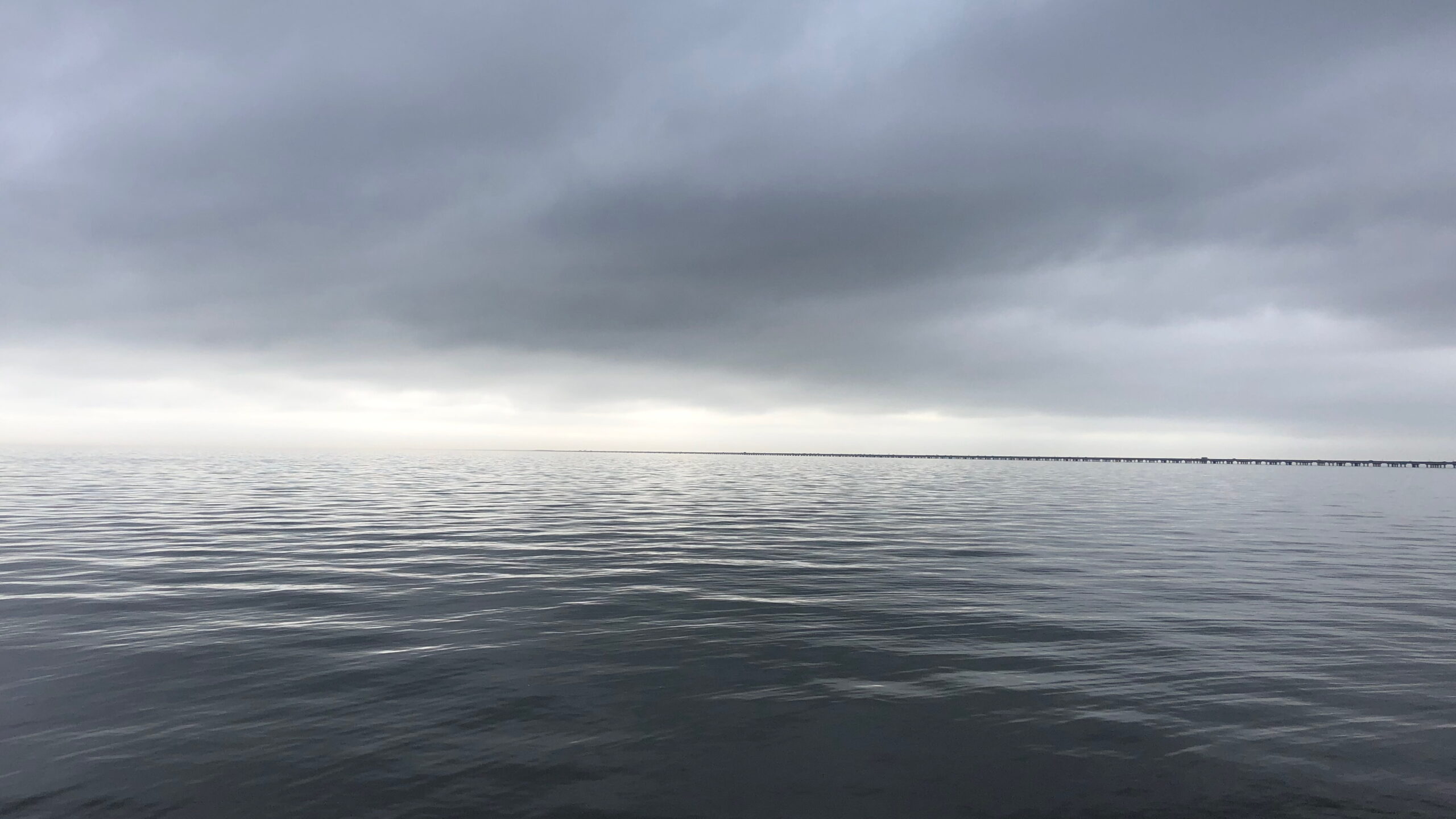 A wide expanse of calm water under a dark, overcast sky. A long, narrow bridge stretches across the horizon in the distance. The scene is moody and serene, with soft ripples on the surface of the water.