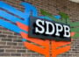 SDPB sign mounted on a brick wall, with bold white letters on a black background, set against a colorful abstract backdrop resembling stylized wings in blue, orange, and green.