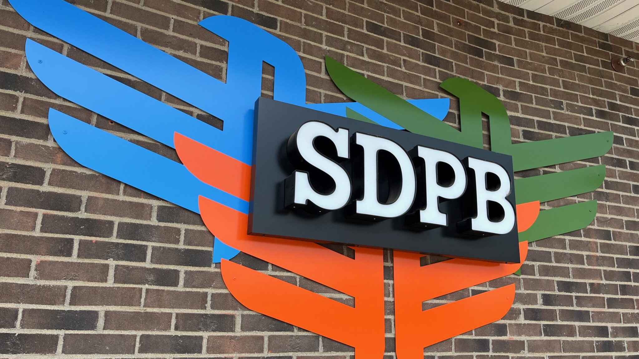 SDPB sign mounted on a brick wall, with bold white letters on a black background, set against a colorful abstract backdrop resembling stylized wings in blue, orange, and green.