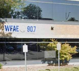 Exterior of WFAE 90.7’s office building with a sign displaying the station’s call letters and frequency. The building has large reflective windows showing trees, parked cars, and a couple of people walking nearby. Several parking signs are visible in the foreground.