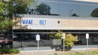 Exterior of WFAE 90.7’s office building with a sign displaying the station’s call letters and frequency. The building has large reflective windows showing trees, parked cars, and a couple of people walking nearby. Several parking signs are visible in the foreground.