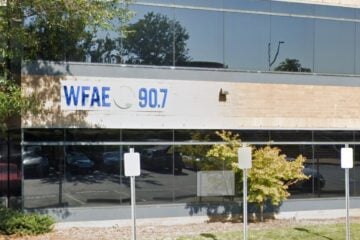 Exterior of WFAE 90.7’s office building with a sign displaying the station’s call letters and frequency. The building has large reflective windows showing trees, parked cars, and a couple of people walking nearby. Several parking signs are visible in the foreground.