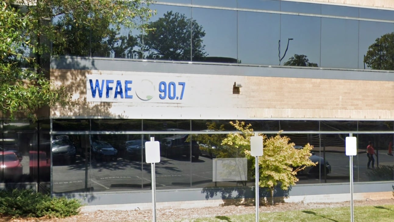 Exterior of WFAE 90.7’s office building with a sign displaying the station’s call letters and frequency. The building has large reflective windows showing trees, parked cars, and a couple of people walking nearby. Several parking signs are visible in the foreground.