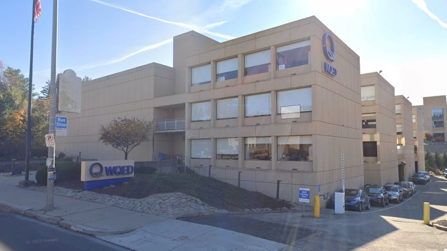 he exterior of WQED’s headquarters in Pittsburgh, a beige modernist building with large rectangular windows and the WQED logo on two corners. A sign with the WQED logo sits near a bus stop and flagpole on the sidewalk in front. Several cars are parked along the building’s driveway.