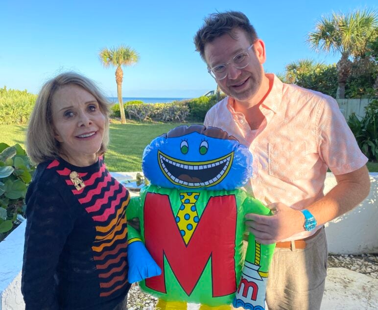 Elayne Reiss-Weimann and Jason Collum pose with a Letter People large inflatable character shaped like the letter M, decorated with a colorful suit and tie. Palm trees and the ocean are visible in the background.